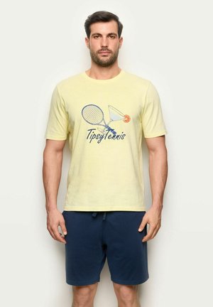 Hombre vistiendo una camiseta amarilla "Tipsy Tennis" con un gráfico de raqueta de tenis y cóctel, pantalones cortos azul marino, de pie contra un fondo blanco.