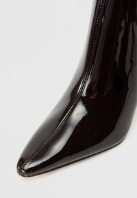 Bota de bico fino em pele patent preta brilhante, com um silhueta elegante e uma textura suave, apresentando um fecho de zíper na lateral.