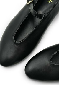 Bailarinas de cuero negro con una textura suave, punta redondeada y un corte distintivo. Cuentan con una banda delgada con un detalle de hebilla dorada.