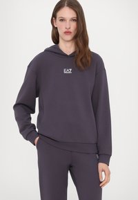 Mörklila hoodie i mjukt material, med en framficka, ribbade muddar och en broderad vit logotyp på bröstet. Matchas med byxor i samma färg.