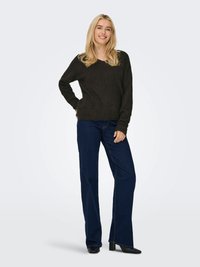 Plavuša koja se smiješi, nosi tamnosmeđi džemper, plave traperice i crne čizme s potpeticama, stoji s jednom rukom na bokovima ispred jednobojne pozadine.