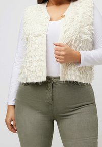 Cremefarbene Weste mit flauschigem, strukturiertem Stoff und Knopfverschluss, kombiniert mit hellgrünen, eng anliegenden Hosen. Darunter ein langärmliges weißes Oberteil.