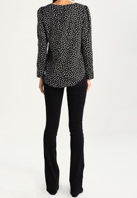 Schwarze Bluse mit langen Ärmeln und einem weißen Punktmuster, mit gesammelten Ärmeln und einem Reißverschluss im Rücken. Kombiniert mit schwarzen Schlaghosen.