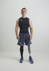 Idrottare klädd i svart, åtsittande ärmlös topp, marinblå shorts med mesh-detaljer och en protes i benet. Har blåa sportskor.