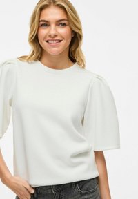 Haut blanc à manches courtes avec un col rond et des manches bouffantes, fabriqué dans un tissu léger et lisse. Associé à un pantalon en denim foncé.