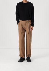 Svart långärmad skjorta med ribbad textur, beige veckade byxor i avslappnad passform och svarta klumpiga skor mot en neutral bakgrund.