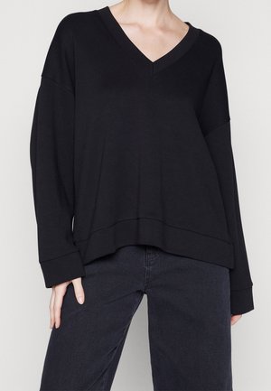 Person trägt einen lockeren schwarzen V-Ausschnitt mit langen Ärmeln und dunkle Hose, steht vor einem einfarbigen hellen Hintergrund.