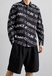 Svart skjorta med knappar som har stor vit text "VERSACE JEANS COUTURE" i olika typsnitt, tillsammans med lösa svarta shorts.