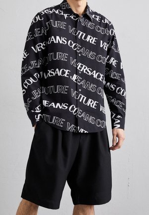 Camisa negra de botones con grande texto blanco "VERSACE JEANS COUTURE" en varias tipografías, combinada con pantalones cortos negros holgados.