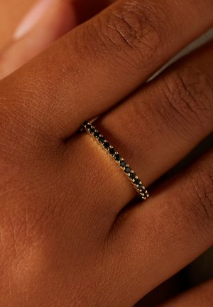 Gouden ring met een smalle band, met bovenaan een rij gelijkmatig verdeelde zwarte stenen. Gestructureerde huidachtergrond.