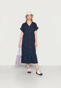 Vestido midi azul marinho com mangas curtas, frente de botões e fendas laterais. Usado com sandálias brancas de slide e uma bolsa de malha roxa.