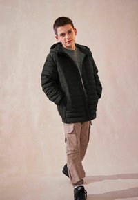 Giacca imbottita nera con zip frontale e cappuccio, abbinata a pantaloni cargo beige chiaro e sneakers nere. Design semplice e pratico.
