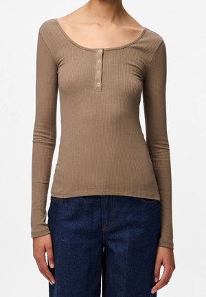Personne portant un haut marron côtelé ajusté à manches longues avec un col boutonné et un jean en denim bleu foncé, debout devant un fond uni.