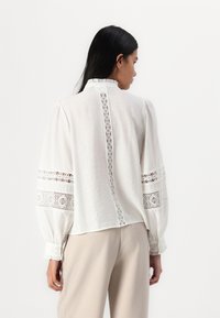 Weiße Baumwollbluse mit Puffärmeln, die mit Spitzenverzierungen an den Ärmeln und am Rücken versehen ist. Strukturiertes Gewebe und ein hoher Kragen runden das Design ab.