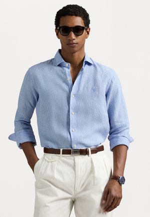 Camicia a maniche lunghe blu chiaro con texture, bottoni sul davanti e polsini arrotolati, abbinata a pantaloni crema e una cintura marrone. Occhiali da sole neri e orologio.