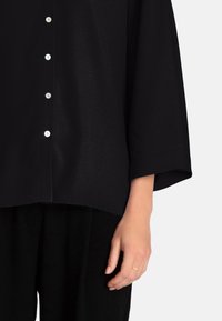 Top nero, di taglio morbido, con una texture liscia, caratterizzato da una fila di bottoni argentati e maniche a tre quarti, abbinato a pantaloni neri.