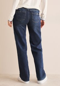 Donkerblauwe denim jeans met een ontspannen pasvorm, voorzien van twee achterzakken en gestikte details, gecombineerd met witte sneakers.