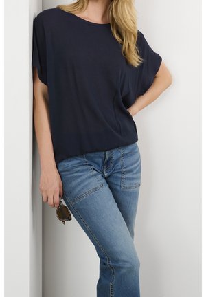 Frau lehnt an einer Wand, trägt ein lockeres dunkelblaues Hemd und hellblaue Jeans, hält eine Sonnenbrille in einer Hand.