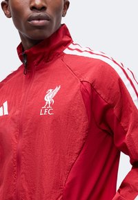 Νεαρός άνδρας που φοράει κόκκινο μπουφάν Liverpool FC με λευκές ρίγες στα μανίκια και το λογότυπο L.F.C. στο στήθος.