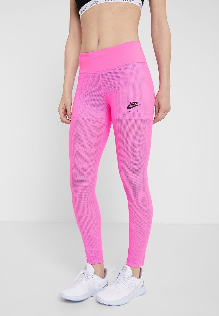 Žena v neonově růžových legínách Nike Air a bílých běžeckých botách Nike, stojící proti jednoduchému bílému pozadí.