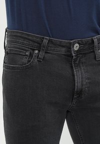 Vaqueros de mezclilla negra con corte ajustado. Presentan un diseño de cinco bolsillos, botones plateados y costuras visibles sobre una tela texturizada.