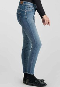 Jeans skinny en denim bleu moyen avec un léger délavage, cinq poches et une étiquette en cuir avec logo à la taille. Portés avec des chaussures noires.