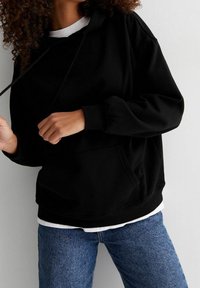 Sudadera negra con capucha y un gran bolsillo frontal, con una textura suave y un corte holgado, superpuesta sobre una camisa blanca y combinada con jeans azules.