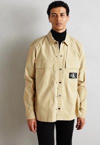 Beige Hemd mit Knöpfen, zwei vorderen Taschen, einem Logo-Patch auf der linken Seite und einem Kragen. Über einem schwarzen Rollkragenpullover getragen.
