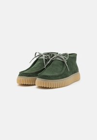 Scarpe alla caviglia in suede verde con una tomaia testurizzata, caratterizzate da lacci grigi a contrasto e una suola in gomma traslucida chunky.