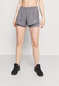 Pantalones cortos deportivos grises con cintura elástica, forro interior y un logotipo blanco de Nike en el lado izquierdo. Se llevan con zapatillas negras.