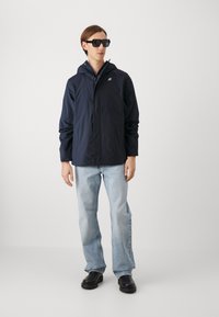 Giacca impermeabile blu navy con cappuccio, maniche lunghe e logo; abbinata a jeans azzurri chiari e scarpe nere spesse, modello che indossa occhiali da sole.