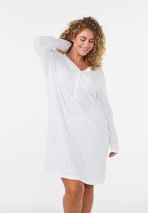 Vrouw met krullend haar die een witte jurk met lange mouwen draagt en glimlacht, met één hand haar nek aanraking tegen een effen witte achtergrond.