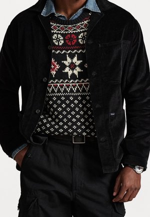 Homme portant une veste en velours côtelé noire par-dessus un pull en tricot à motifs rouges, blancs et noirs, pantalon sombre et une montre-bracelet.