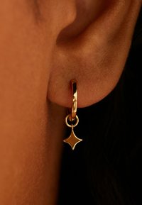 Boucle d'oreille en forme d'anneau doré avec un pendentif brillant en forme d'étoile. Surface lisse et finition polie. Suspendue à une boucle circulaire simple.