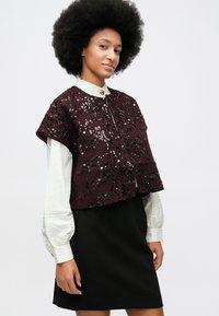 Ženska z afro pričesko, oblečena v belo bluzo z dolgimi rokavi in gumbi na manšetah, kratko jakno z maroon bleščicami in črno krilo, stoji proti beli ozadju.