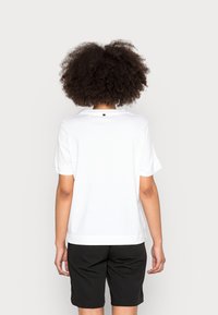 Camiseta blanca de manga corta hecha de algodón, con un corte relajado y un pequeño detalle en negro en la parte trasera. Combinada con pantalones cortos negros.