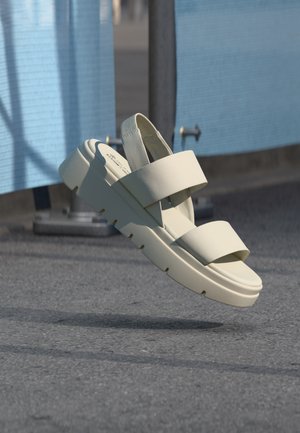 Εκρού πλατφόρμα σανδάλι με δύο φαρδιές λουριά και λουράκι πίσω από τη φτέρνα, αιωρούμενο πάνω από επιφάνεια από άσφαλτο με μπλε φράγμα στο βάθος.