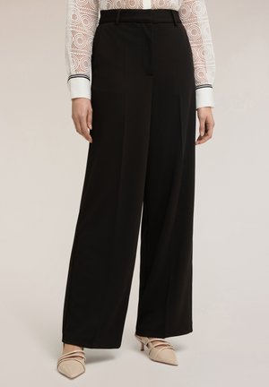 Donna che indossa pantaloni neri a gamba larga, una camicetta bianca in pizzo con motivi circolari e tacchi a punta beige con cinturini.