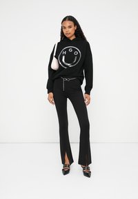 Zwarte hoodie met een zilveren logo, gecombineerd met zwarte flared broek met sleuven. Geaccessoireerd met een kleine roze tas en zwarte hoge hakken.