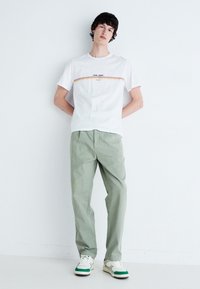 Biała koszulka z krótkim rękawem z wielokolorowym poziomym paskiem i logo, zestawiona z jasnymi zielonymi chinosami i białymi trampkami z zielonymi akcentami.