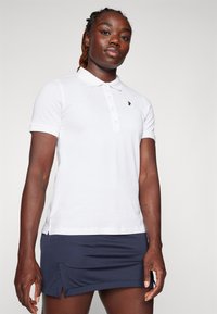 Weißes Poloshirt mit kurzen Ärmeln und einem Drei-Knopf-Verschluss, mit einem kleinen schwarzen Logo auf der Brust. Getragen mit einem dunkelblauen Rock.