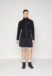 Črna flis jakna z visokim ovratnikom, zadrgo in logotipom. Združena s temnimi kratkimi hlačami z velikimi žepi in črnimi pohodniškimi čevlji.