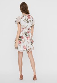 Vaatteet kukkakuosisessa kevyessä, läpikuultavassa kankaassa, jossa on vaaleanpunaisia ja vihreitä kukkia valkoisella taustalla. Mekossa on kerrokselliset röyhelöt ja lyhyet hihat.