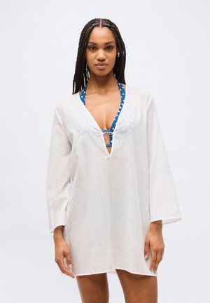 Femme aux cheveux tressés portant un paréo de plage blanc à manches longues et col en V, sur un haut de bikini halter bleu à pois, sur un fond uni.