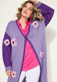 Lång, öppen cardigan i lila stickad med rosa och lilafärgade blommorapplikationer. Kombinerad med en rosa skjorta, vita byxor och ringörhängen.