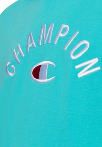 Sudadera de algodón azul claro con bordado en blanco de "CHAMPION" y un logo circular en acentos rojos y blancos. Tela texturizada.