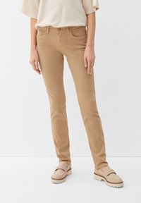 Osoba w beżowych dopasowanych spodniach i jasnobeżowych loafersach stoi na tle jednolitego białego tła.