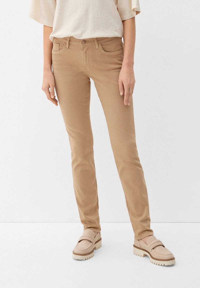 Osoba w beżowych dopasowanych spodniach i jasnobeżowych loafersach stoi na tle jednolitego białego tła.