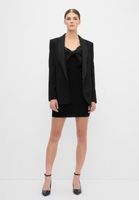 Černé přiléhavé šaty s mašlí na výstřihu, doplněné o oversized černý pruhovaný blazer a černé vysoké podpatky.