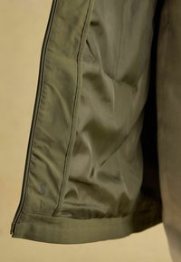 Jachetă verde măsliniu la interior, cu o textură netedă, umplutură matlasată și fermoar. Are un logo aproape de tiv.